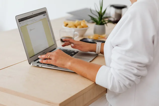 Close-up of a woman typing on a laptop at a home office desk with coffee and snacks. Telearbeit in der Schweiz: Vereinbarung, Ausrüstung und Kosten