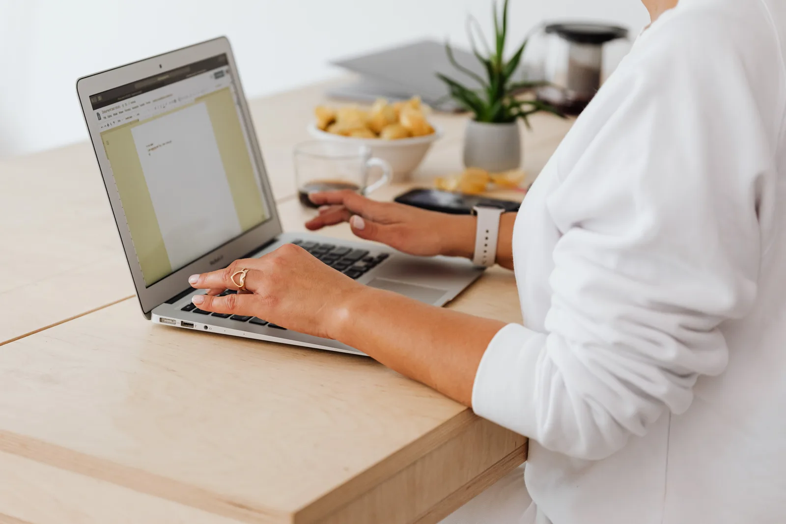 Close-up of a woman typing on a laptop at a home office desk with coffee and snacks.